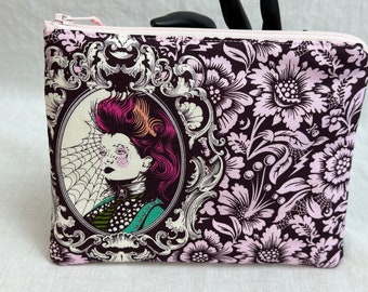 May include: A zippered pouch with a dark purple floral pattern and a pink zipper. The pouch features a framed portrait of a woman with pink hair and a spiderweb design. The overall theme is gothic and vintage.