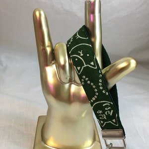 Puede incluir: Un cordón de muñeca de tela verde con un patrón de cara de gato blanco y texto japonés. El cordón tiene una correa verde oscuro y un cierre de metal plateado con un anillo partido.