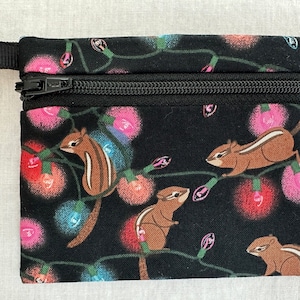May include: A small, black zippered pouch featuring a repeating pattern of brown chipmunks and colorful string lights. The pouch has a black zipper and a small black loop on one side.