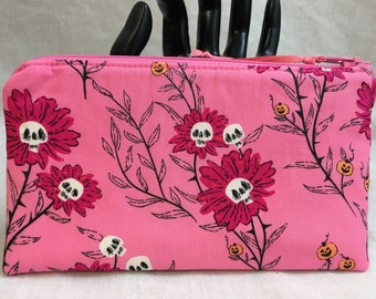 May include: Pink zippered pouch with a floral print featuring black stems, leaves, and pink flowers with white skull faces.  The background is pink with orange jack-o-lantern faces scattered throughout.