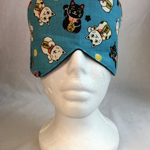 May include: A blue sleep mask with a pattern of white and black lucky cats. The cats are holding up a paw and have a gold coin on their head.