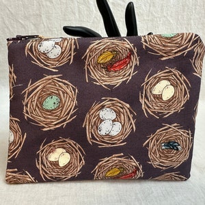 May include: A brown zippered pouch with a repeating pattern of birds' nests with eggs. The nests are made of twigs and the eggs are white, green, and yellow.