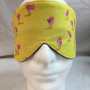 May include: A yellow sleep mask with a repeating pattern of pink birds. The mask is made of fabric and has a black interior lining. The mask is designed to block out light.