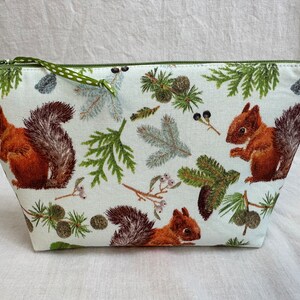 May include: A white zippered pouch with a green zipper pull. The pouch is decorated with a repeating pattern of brown squirrels, green pine branches, and brown pine cones.