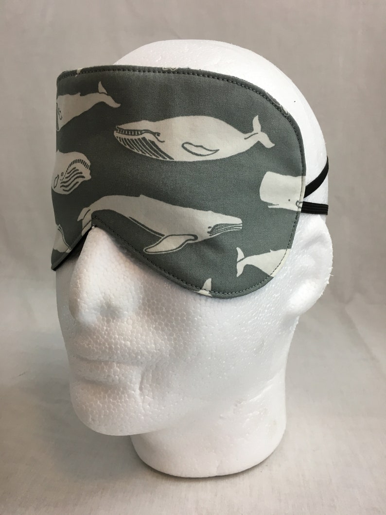Puede incluir: Una m&aacute;scara para dormir gris con un patr&oacute;n de ballena blanca. La m&aacute;scara tiene una banda el&aacute;stica para sujetarla a la cabeza.