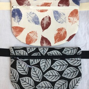 May include: Two fabric fanny packs with leaf patterns. The top fanny pack is cream with blue and brown leaves. The bottom fanny pack is black with white leaves.