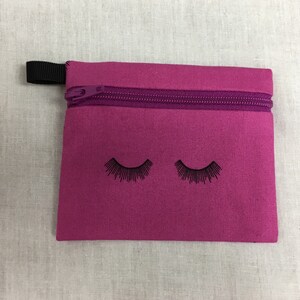 Könnte beinhalten: Eine kleine rosa Reißverschlusstasche mit einem schwarzen Reißverschluss und zwei schwarzen Wimpern, die auf der Vorderseite aufgedruckt sind.