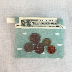May include: A small, teal blue zippered pouch with white polka dots. The pouch is open and contains a one dollar bill and five coins. The coins are a quarter, a nickel, a dime, and two pennies.