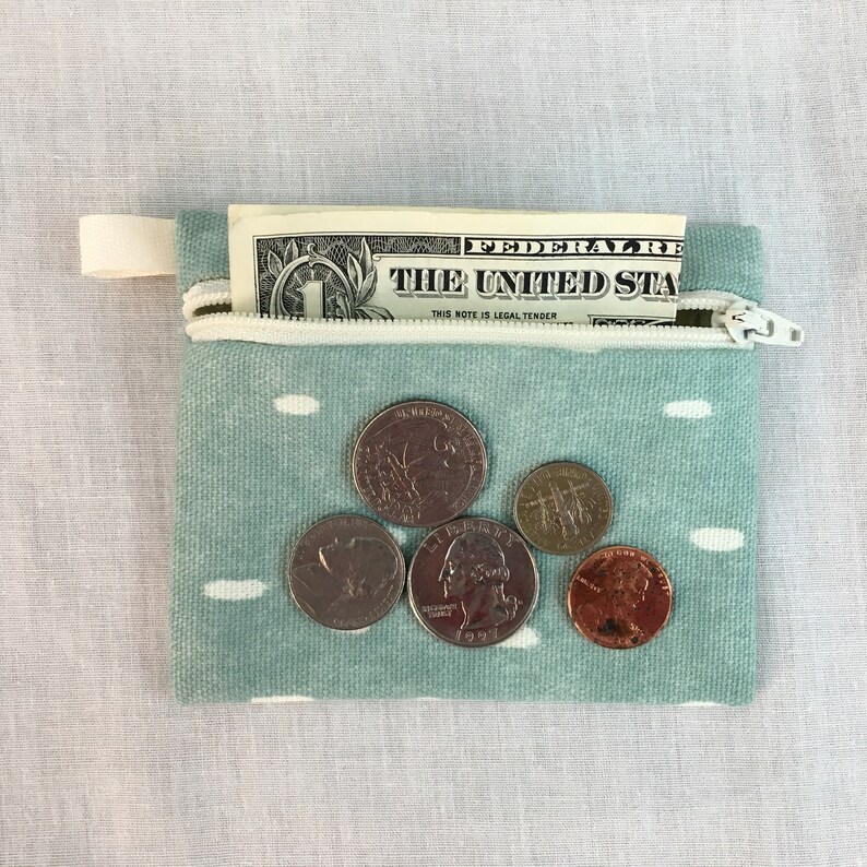 May include: A small, teal blue zippered pouch with white polka dots. The pouch is open and contains a one dollar bill and five coins. The coins are a quarter, a nickel, a dime, and two pennies.