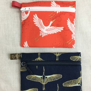 May include: Two small zippered pouches with a crane print. The top pouch is red with white cranes and gold accents. The bottom pouch is navy blue with gold cranes.
