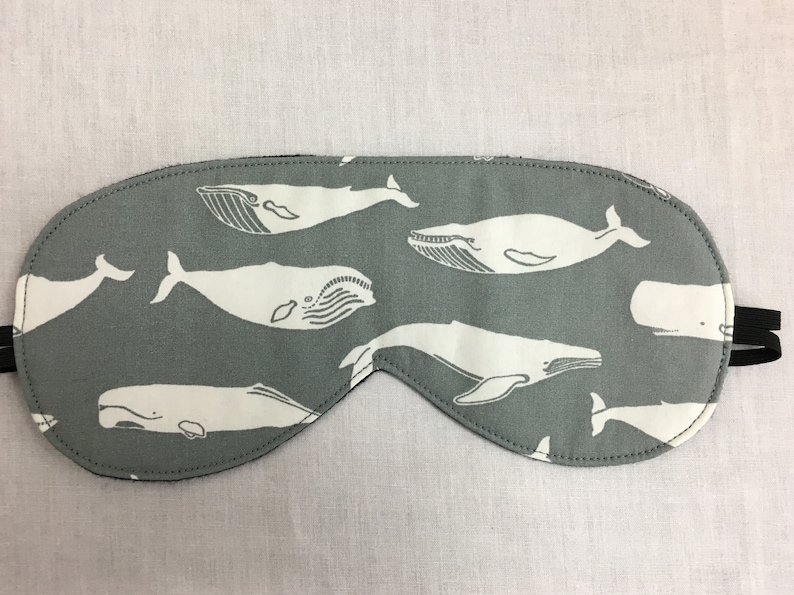 Puede incluir: Una m&aacute;scara para dormir gris con un patr&oacute;n de ballena blanca. La m&aacute;scara est&aacute; hecha de tela suave y tiene una correa el&aacute;stica.
