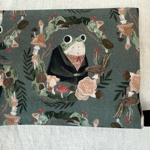 May include: A green fabric pouch with a black zipper pull. The pouch features a repeating pattern of a frog wearing a black coat, mushrooms, and leaves on a green background.