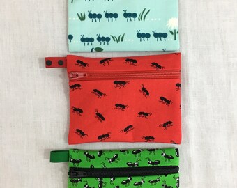 May include: Three zippered pouches with a repeating pattern of black ants on different coloured backgrounds. The top pouch is light blue with a white daisy and a red strawberry. The middle pouch is red. The bottom pouch is green.