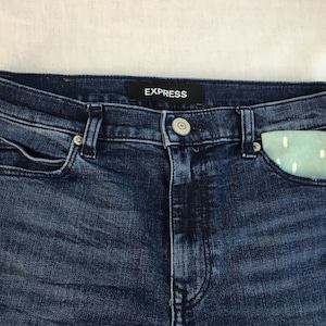 May include: Dark blue denim jeans with a small, light blue and white patterned pouch in the right front pocket. The jeans have a black label with the word "EXPRESS" on the waistband.