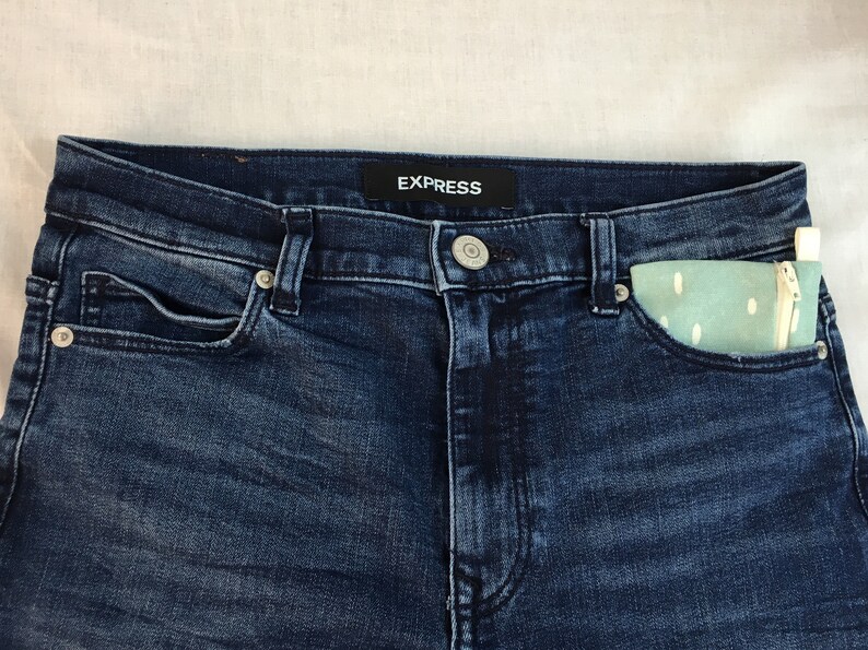 May include: Dark blue denim jeans with a small, light blue and white patterned pouch in the right front pocket. The jeans have a black label with the word "EXPRESS" on the waistband.