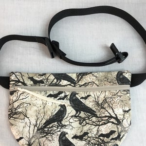 May include: A black and white fanny pack with a zipper closure. The fabric features a pattern of ravens and bats on a beige background.