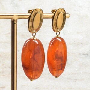 Könnte beinhalten: Ein Paar goldfarbene Ohrringe mit ovalen Oberteilen und großen, bernsteinfarbenen, ovalen Tropfen. Die Ohrringe werden auf einem goldenen Ständer vor einem weißen Marmorhintergrund präsentiert. Die Tropfen haben einen Marmoreffekt.