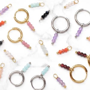 Op de afbeelding: Een assortiment oorringen met kleurrijke kralenbedels. De oorbellen hebben gouden en zilveren ringen met bungelende bedels in verschillende kleuren, waaronder roze, paars en blauw. De kralen zijn klein en rond, en de oorbellen worden gepresenteerd op een wit marmeren oppervlak.