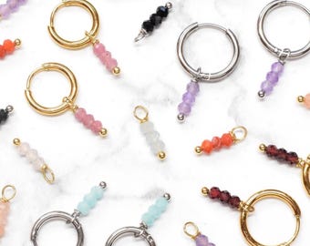 Puede incluir: Una colección de pendientes de aro dorados y plateados y dijes individuales con pequeñas cuentas de piedras preciosas facetadas y multicolores. El surtido incluye varios colores como rosa, morado, rojo, negro, azul claro y blanco.