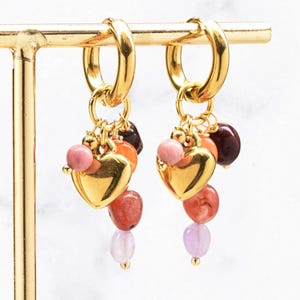 Op de afbeelding: Een paar gouden oorringen met bungelende bedels. Elke oorbel bevat een gepolijste gouden hartbedel omringd door kleine, onregelmatige kralen in tinten roze, oranje, rood en donkerbruin, allemaal hangend aan een ronde gouden oorring.