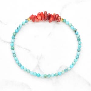Op de afbeelding: Een kralenarmband bestaande uit een cirkelvormige streng van kleine, ronde, lichtblauwe en turquoise kralen. Het bovenste gedeelte van de armband is geaccentueerd met een cluster van onregelmatige, rode, koraalkleurige steensplinters, geflankeerd door twee kleine goudkleurige metalen kralen.