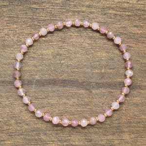 Può includere: Un braccialetto circolare di perline composto da piccole pietre preziose sfaccettate di colore rosa chiaro, alternate a minuscole perline distanziatrici metalliche color oro. Il braccialetto è disposto in un semplice cerchio continuo.