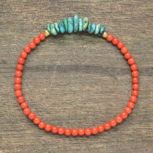 Könnte beinhalten: Ein kreisförmiges Armband aus kleinen, gleichmäßigen orangefarbenen Perlen und einer zentralen Ansammlung unregelmäßiger, roher türkisfarbener Stein-Chips. Das Armband hat zwei kleine, gerippte goldfarbene Metallabstandshalter zwischen den Perlen.