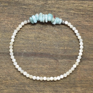 May include: A circular beaded bracelet featuring a long strand of small, round, off-white beads. At the top center, a cluster of irregular, light blue, tumbled stone chips is flanked by two small, metallic silver-toned spacer beads.