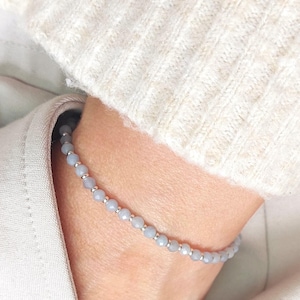 Könnte beinhalten: Ein hellblaues Perlenarmband mit kleinen silbernen Perlen. Das Armband wird am Handgelenk getragen, im Hintergrund ein cremefarbener Pullover. Das Armband besteht aus kleinen, runden, hellblauen Perlen, die mit silbernen Perlen aufgefädelt sind.