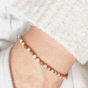 Puede incluir: Una delicada pulsera de cuentas con una mezcla de pequeñas cuentas redondas y facetadas en tonos crema, melocotón y marrón. La pulsera está adornada con pequeñas cuentas doradas. La pulsera se lleva en la muñeca, sobre un fondo de un suéter blanco.