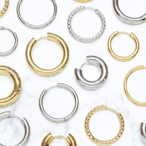 May include: A collection of various hoop earrings in gold and silver tones, featuring both smooth polished surfaces and textured, patterned finishes. The assortment includes different sizes and thicknesses, all designed as circular hoops with hinged posts.