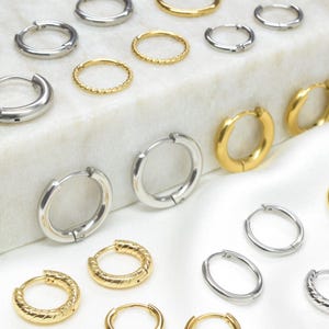 May include: A collection of gold and silver hoop earrings in different styles. Some are plain, others textured or beaded. The earrings are arranged on a white surface, highlighting the variety of designs and sizes, suitable for different tastes.