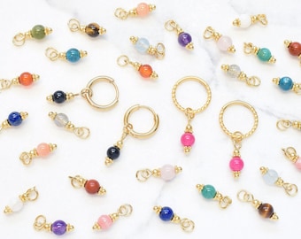 Puede incluir: Una colección de componentes de joyería en tono dorado que incluye varios pendientes de aro pequeños y dijes individuales. Cada pieza presenta una cuenta de piedra preciosa redonda en diversos colores como azul, rosa, verde, morado y marrón, con detalles en metal dorado.