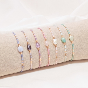 May include: A set of eight delicate bracelets with small beads and a single gemstone or pearl charm in each. The charms are in shades of blue, purple, pink, and white.