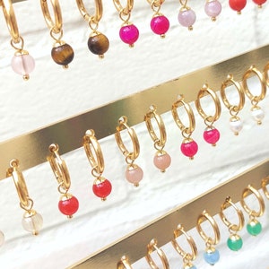 May include: A display of gold hoop earrings with various colored gemstone charms. The charms include pink, red, white, blue, green, and yellow gemstones.