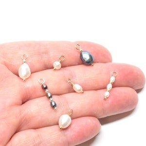 May include: A collection of six pearl and gemstone charms with gold jump rings. The charms include a single black pearl, a single white pearl, a single brown pearl, a single white pearl, a three-pearl strand, and a single white pearl.