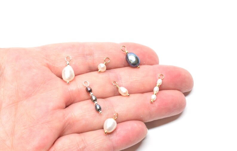May include: A collection of six pearl and gemstone charms with gold jump rings. The charms include a single black pearl, a single white pearl, a single brown pearl, a single white pearl, a three-pearl strand, and a single white pearl.