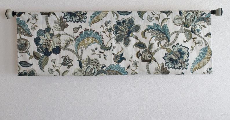 May include: A white valance with a floral pattern in shades of blue, green, and brown. The valance has a rod pocket and is hung on a white wall.