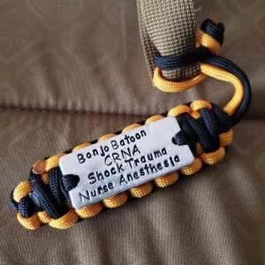 May include: A handmade paracord keychain in black and yellow, with a silver metal tag. The tag is engraved with the words "Bonjo Batoon, CRNA, Shock Trauma, Nurse Anesthesia". The keychain is attached to a tan strap.