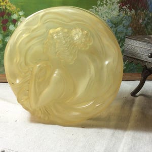 May include: A circular, translucent pale yellow glass powder box lid featuring a molded relief of a figure in profile with flowing hair and draped clothing. The surface has a smooth, frosted texture with raised details depicting the silhouette.
