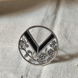 Peut inclure: Une bague argentée avec un motif circulaire. La bague présente un incrustation noire en forme de V et des détails floraux et de vigne. La bague est posée sur un fond en tissu neutre.