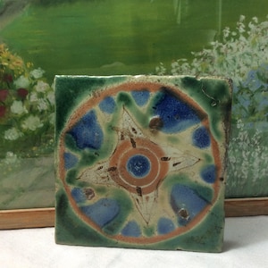 May include: Square ceramic tile with a central design featuring a circle with a blue center, surrounded by an orange ring and blue and green shapes. The tile has a green border and a slightly aged appearance.