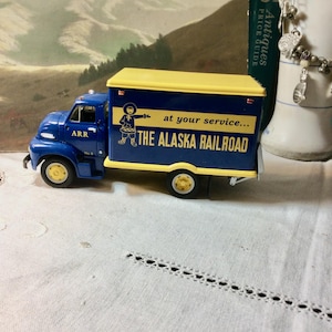 May include: A vintage blue and yellow Alaska Railroad truck model. The truck features the text "at your service... THE ALASKA RAILROAD" on the side. The model has yellow wheels and a yellow top.