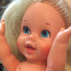 May include: A plastic doll with light skin, bright blue eyes, and an open mouth showing small teeth. The doll has long, light blonde hair and is posed with both arms raised upward, palms facing forward, displaying a cheerful facial expression.