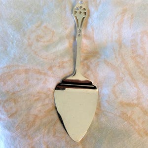 May include: A silver cake server with a decorative handle and a wide, triangular blade. The handle has an openwork design at the top. The server is displayed on a patterned, off-white surface.