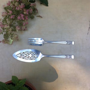 May include: Silver serving fork and pie server with decorative handles and cut-out patterns. The utensils are displayed on a neutral surface, with pink and green foliage in the background. The pie server features an intricate cutout design.