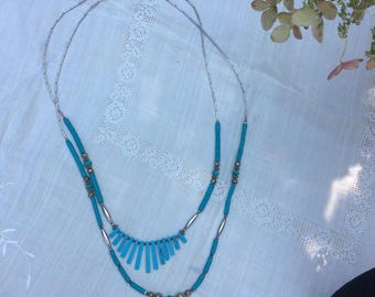 May include: Two-strand necklace featuring turquoise blue beads and silver-tone metal accents. The necklace has a fringe-like design on the lower strand. The jewelry is displayed on a white, textured surface.