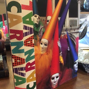 May include: A hardcover book titled ¡CARNAVAL! by Barbara Mauldin. The cover features a photograph of multiple figures wearing white masks and tall, colorful pointed hats in shades of orange, red, blue, and purple, set against a stone architecture.