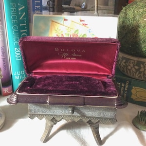 May include: An open, vintage Bulova Fifth Avenue jewelry box. The box is lined with deep red velvet and sits on a decorative metal stand. The Bulova logo is visible inside the box.