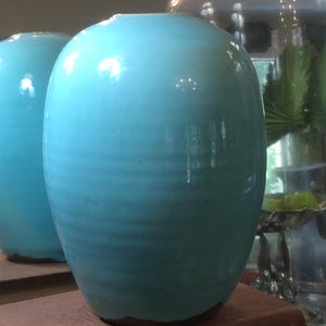 May include: A bright turquoise ceramic vase with a smooth, glossy finish and subtle horizontal ribbed texture. The vase has a rounded, bulbous shape with a narrow opening at the top and a dark, unglazed base that rests on a flat surface.